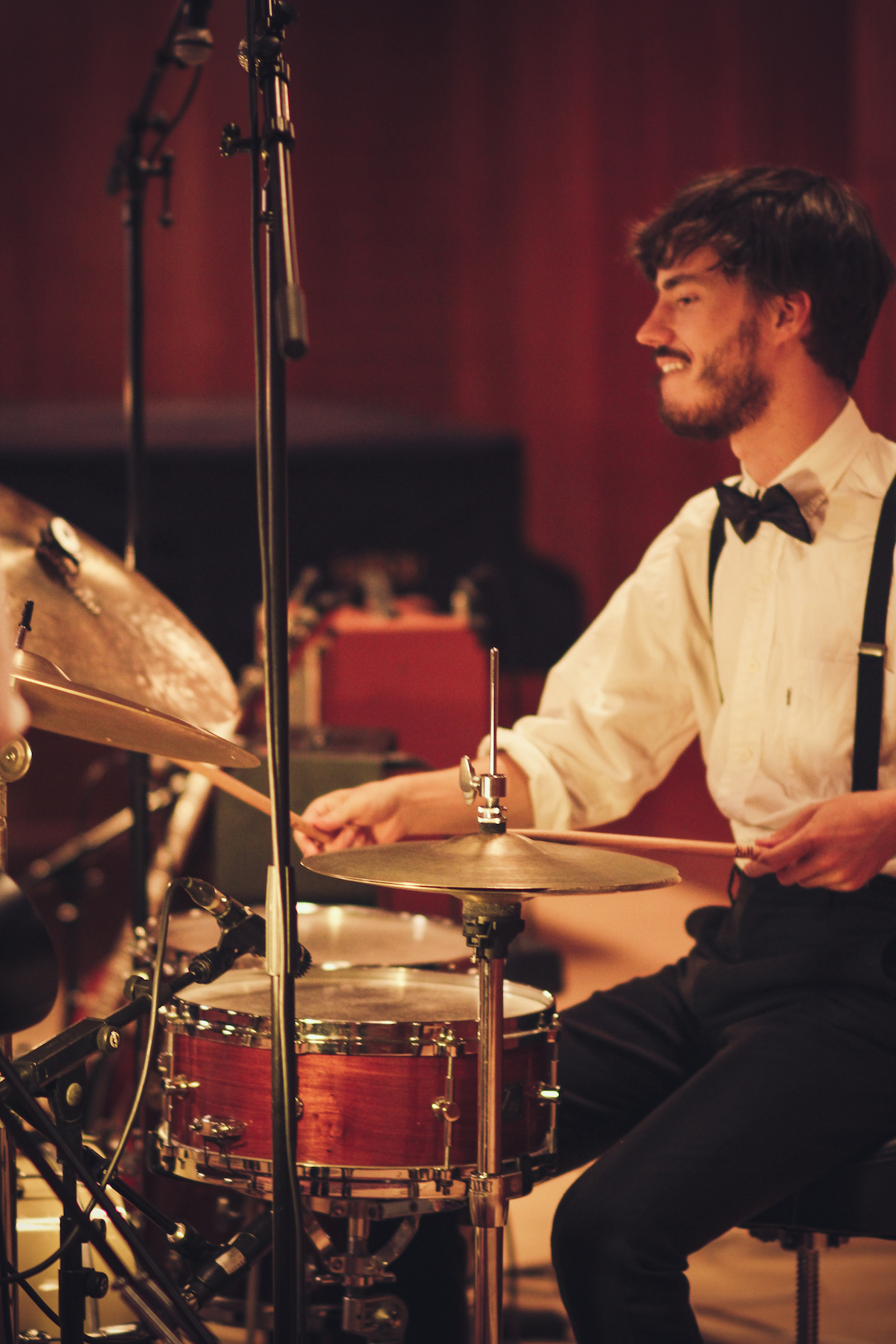 En trommeslager i hvit skjorte og sløyfe spiller på et trommesett omgitt av mikrofonstativer i et innendørs cocktailparty-rom