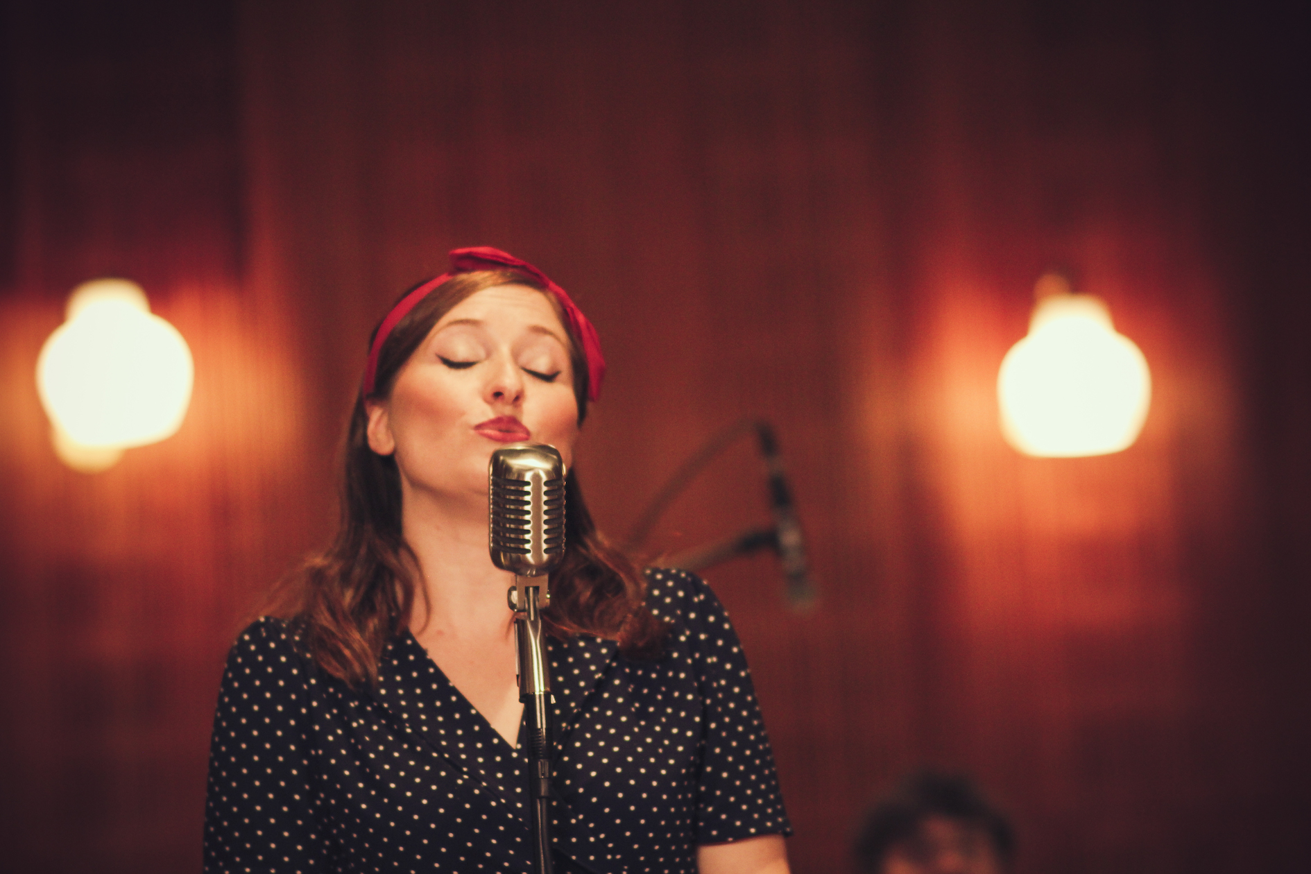 A singer with a red headband stands in front of a microphone and sings, in a warmly lit room