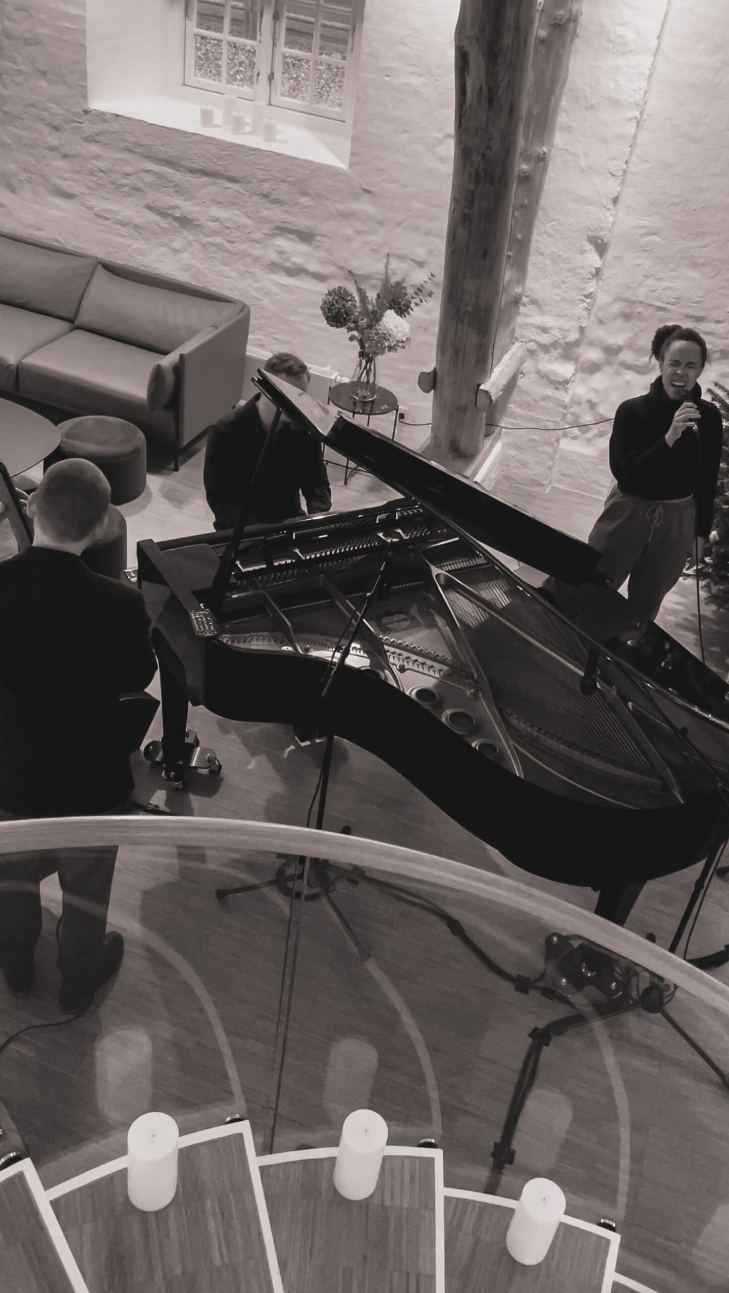 Ein Schwarz-Weiß-Bild zeigt ein Jazz-Trio um einen Flügel in einem gemütlichen Raum mit rustikalen Wänden.