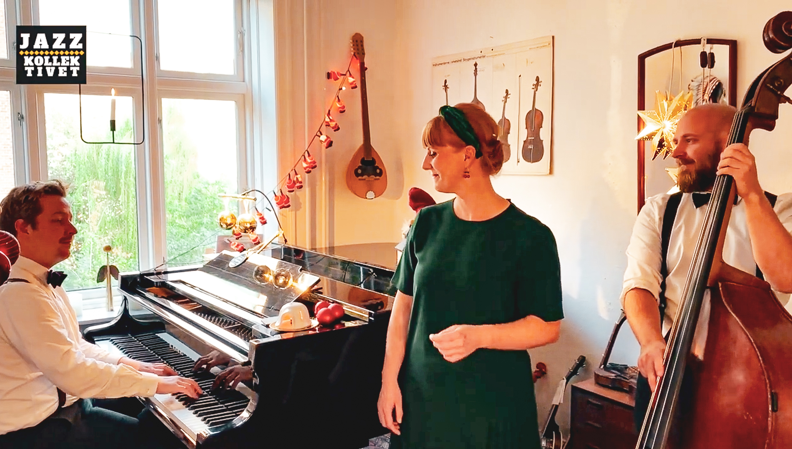 Pianist am Flügel, Sängerin in grünem Kleid und Bassist am Kontrabass vor Weihnachtsschmuck und Wanddekoration im Wohnzimmer