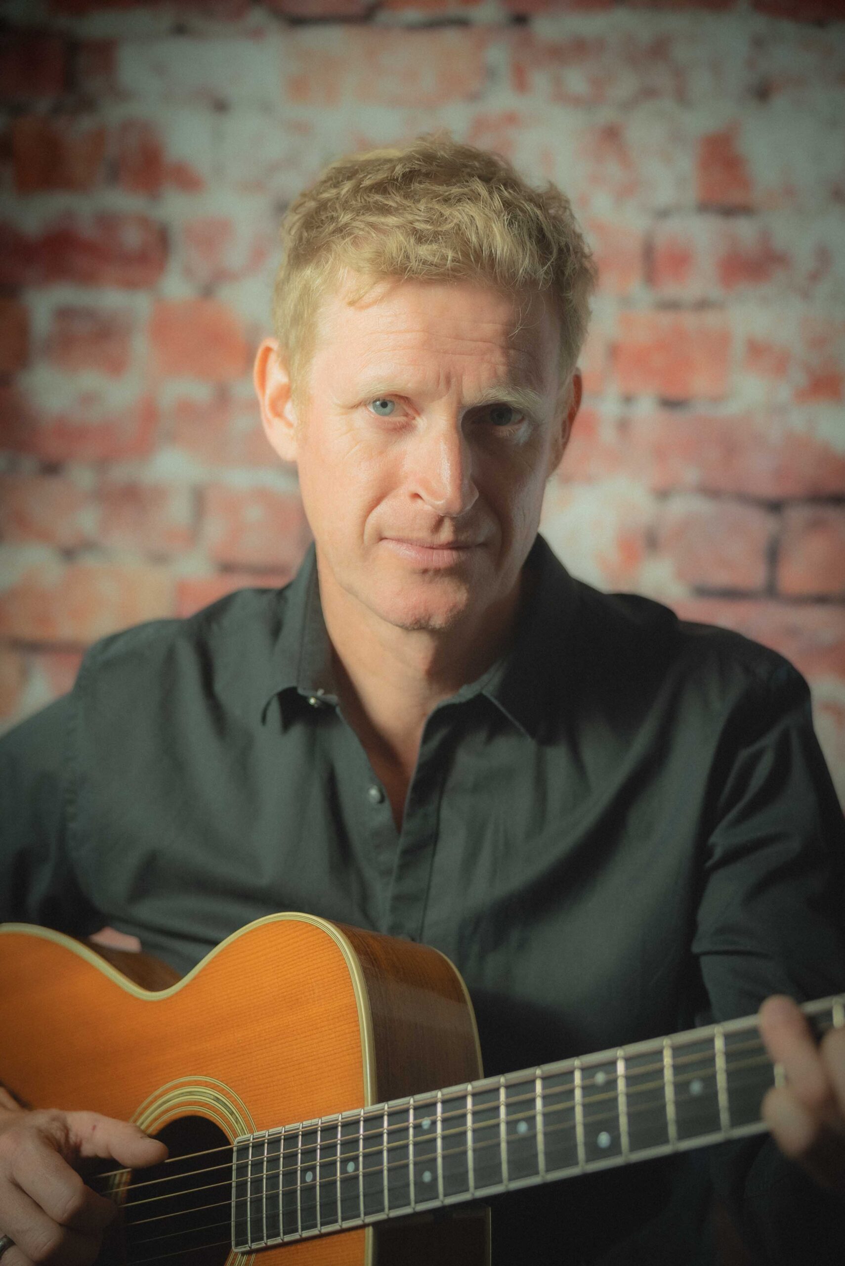 Gitarist som spiller akustisk foran en rå murvegg, med et intenst og fokusert blikk mot kameraet.