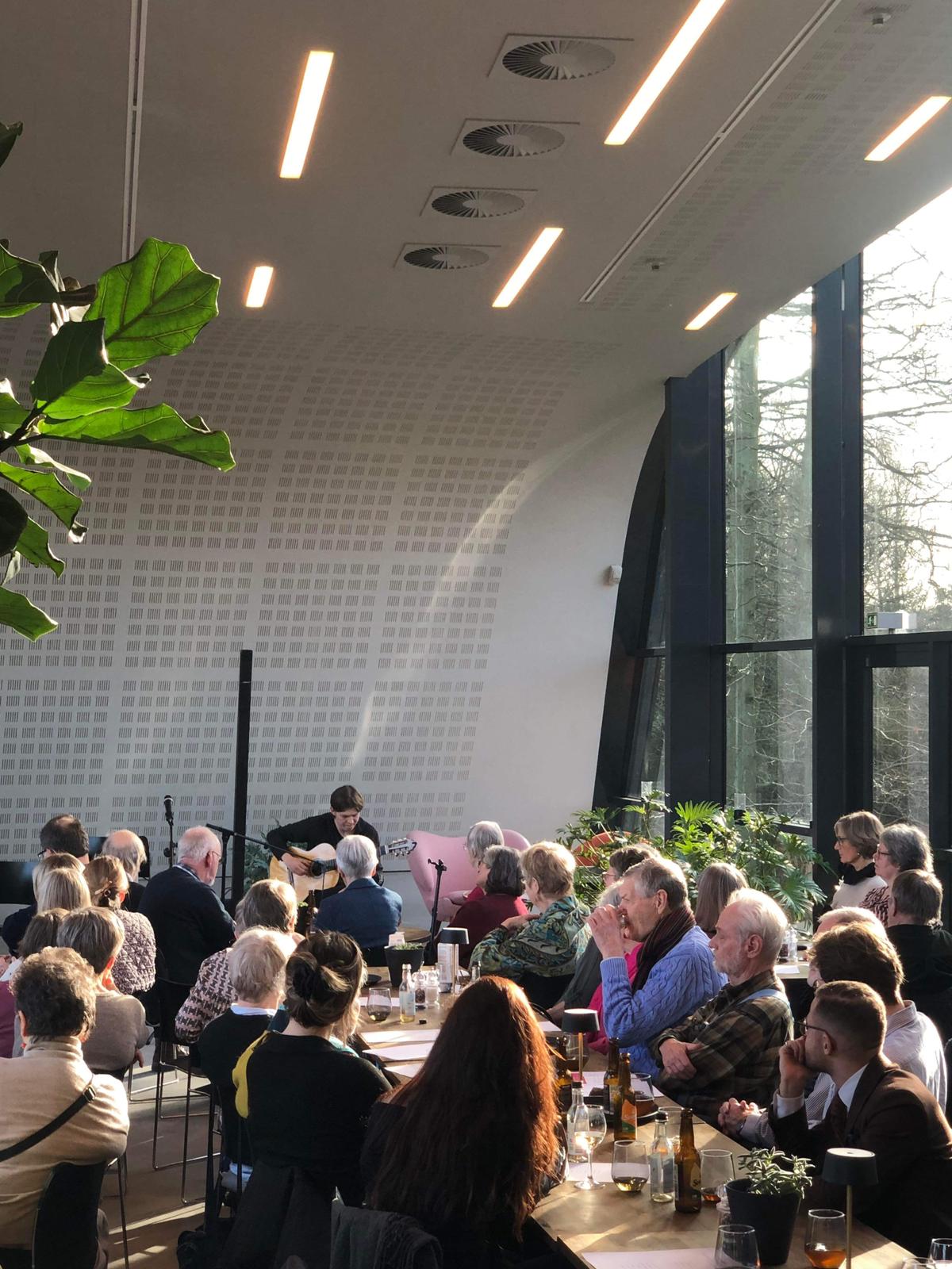 Et publikum lytter intenst til livemusikk til en catwalkshow i et lyst rom der en gitarist spiller solo.