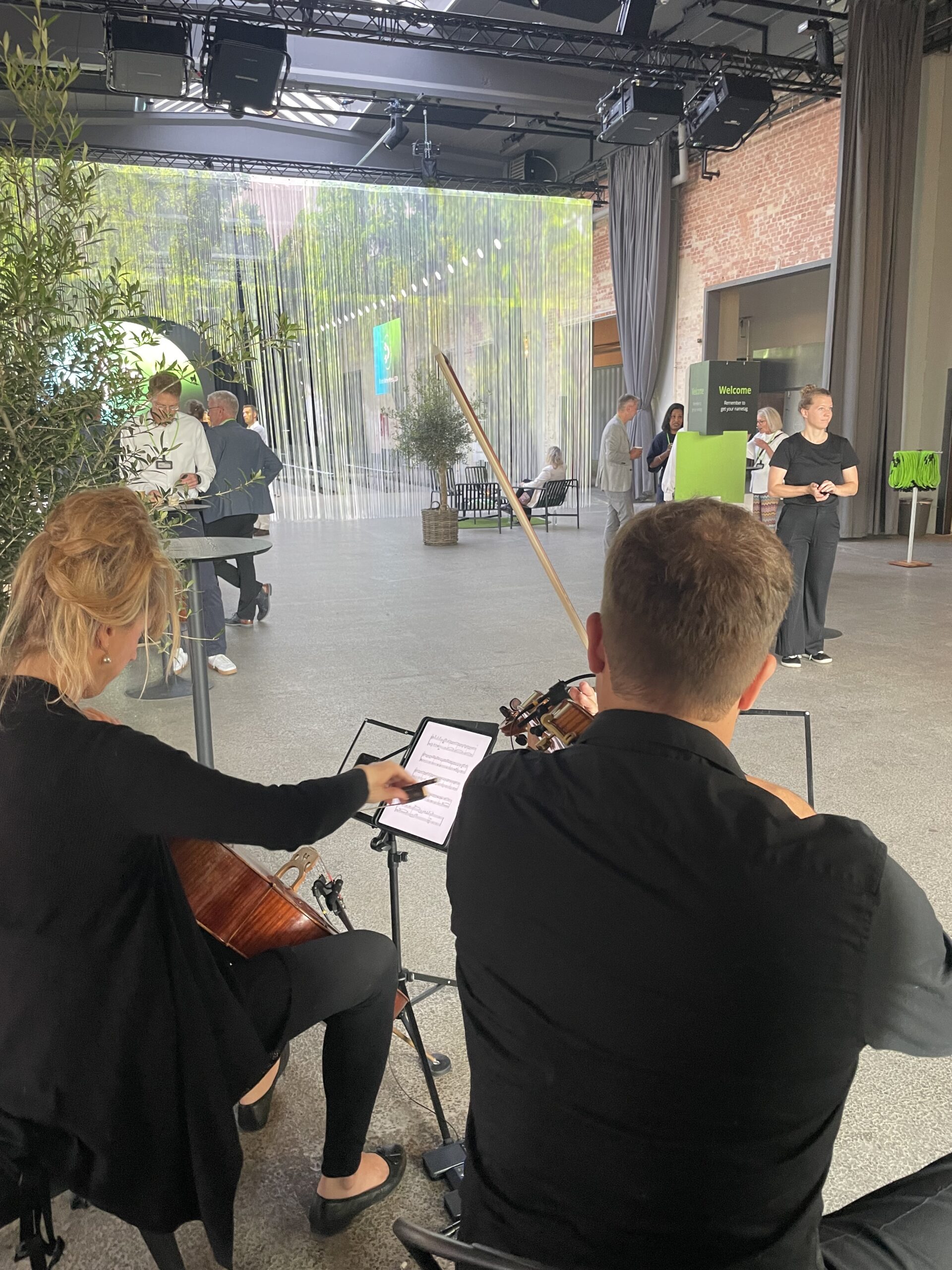 Musikere spiller violin og cello til firmaevent i moderne venue med gæster i baggrunden til reception eller netværksarrangement.