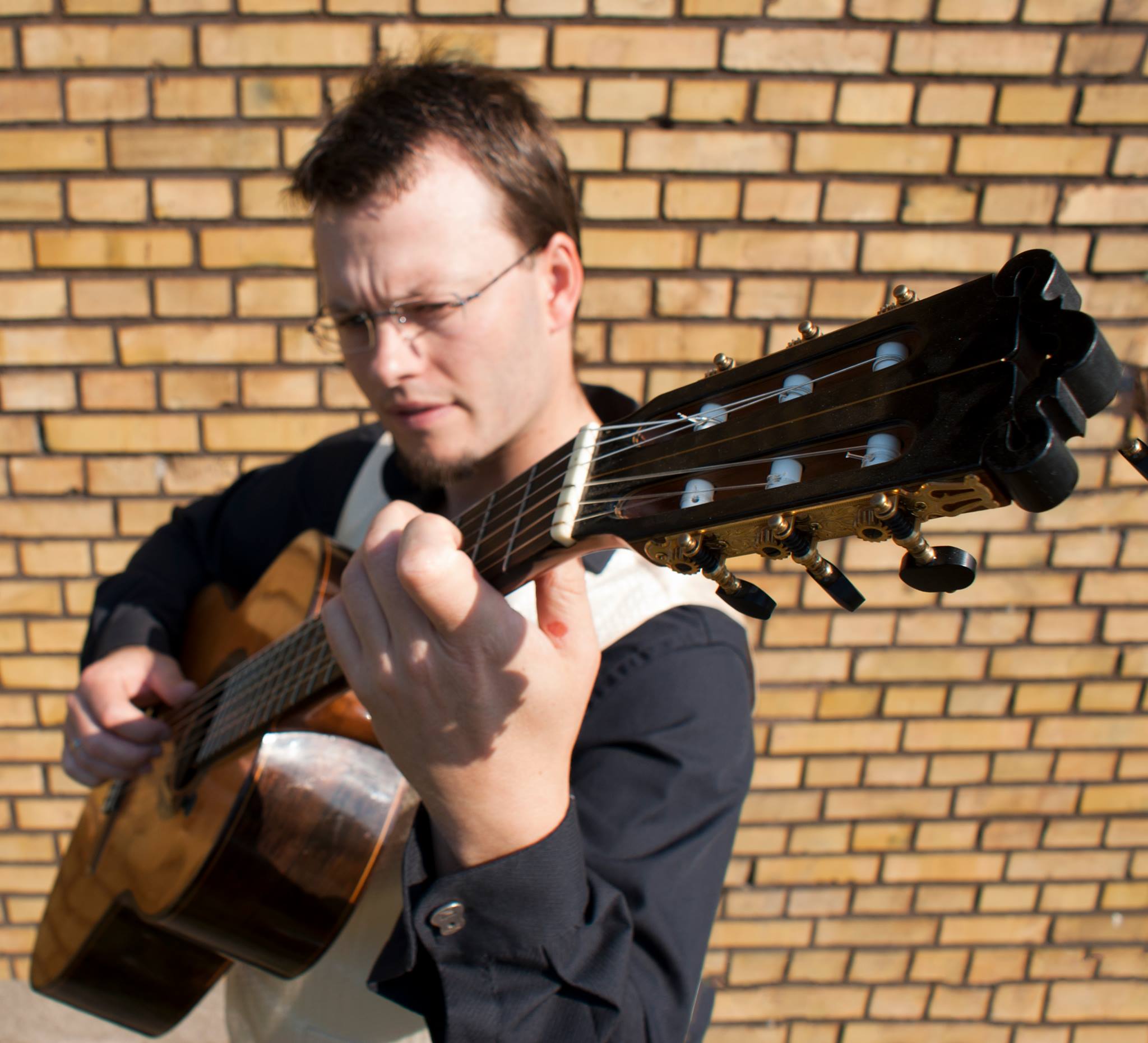 Vor einer Backsteinmauer spielt ein Gitarrist eine Akustikgitarre, während die Kamera den Blickwinkel auf die Saiten und seinen Fokus einfängt.