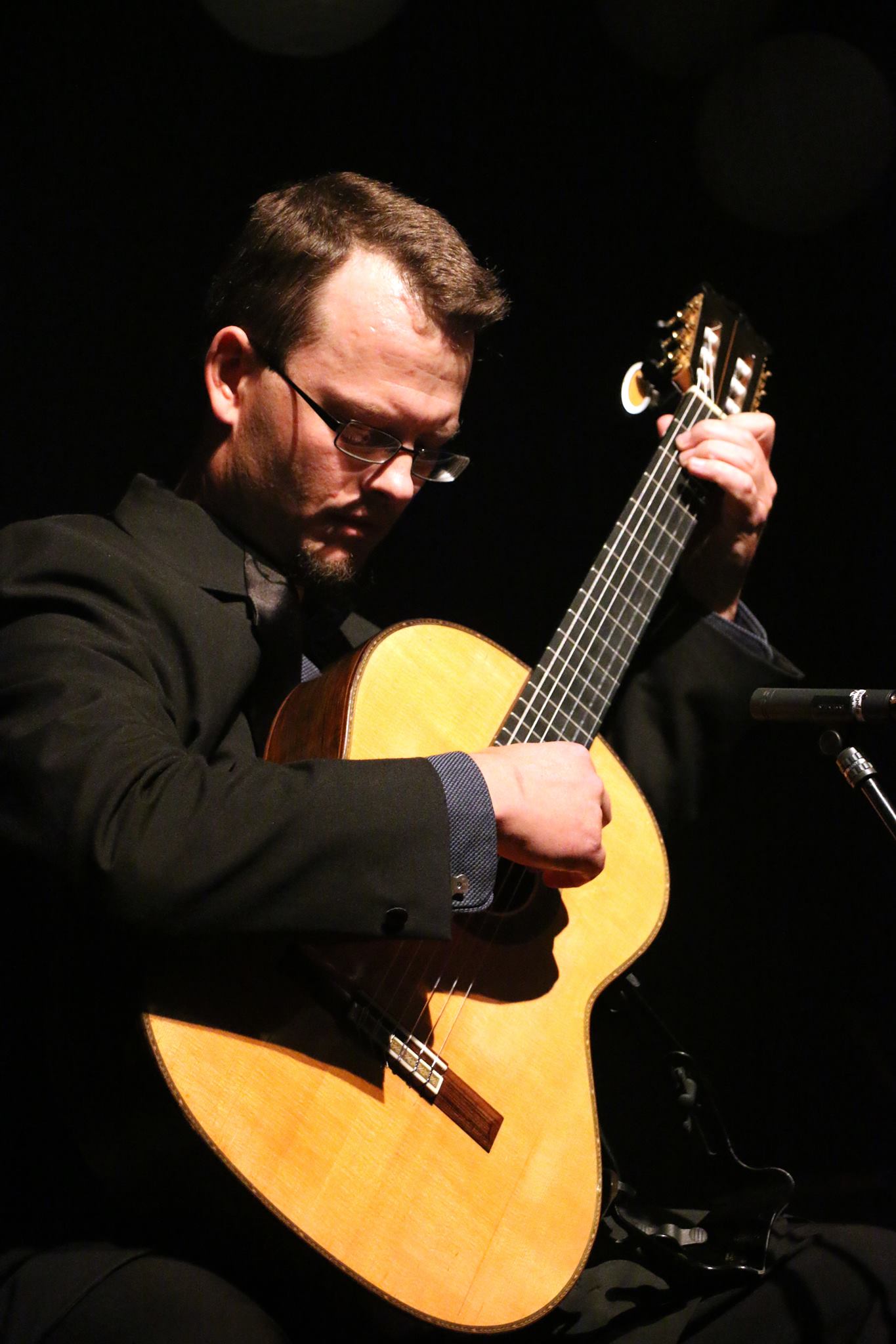 En gitarist spiller konsentrert på sin klassiske gitar i rampelyset, og hans intense fokus gir scenen tilstedeværelse og kraft.