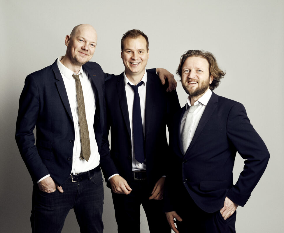 Three men in dark suits standing close together and smiling, exuding professionalism, community and formal music profile