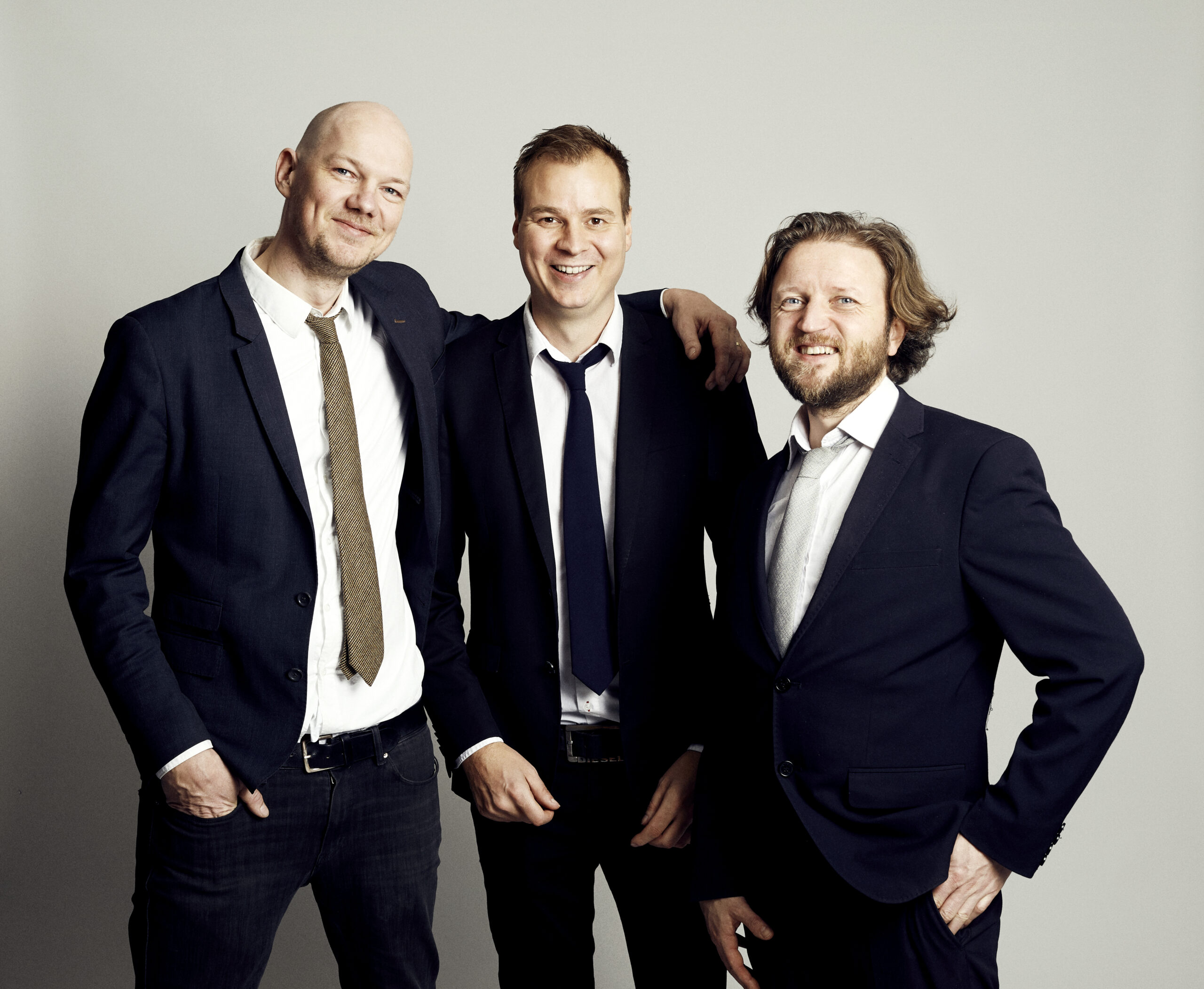 Tre män i mörka kostymer som står tätt tillsammans och ler, utstrålar professionalism, gemenskap och formell musikprofil