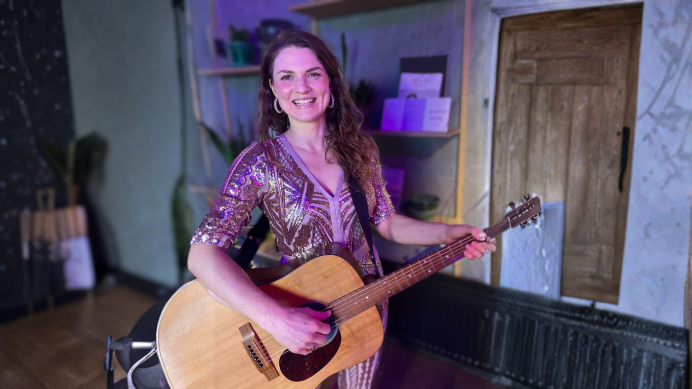 Kvinnlig musiker håller i gitarren i avslappnad inomhusmiljö, vilket signalerar låtskrivande, intimitet och akustisk musik