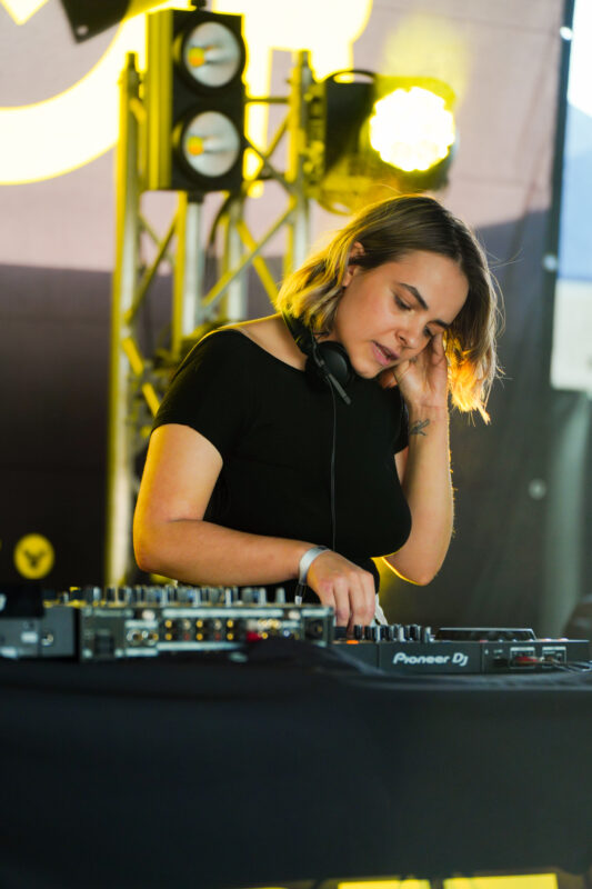 Kvindelig DJ står koncentreret ved DJ-pult med professionelt udstyr, omgivet af scenelys, der signalerer musik, performance og kreativitet