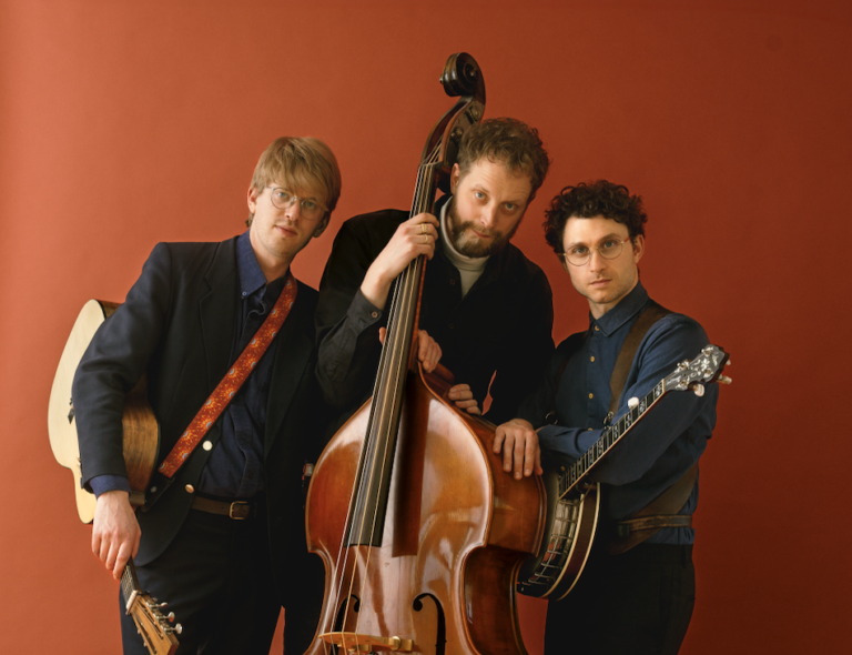 Tre musikere står tett sammen med kontrabass og strykeinstrumenter foran en monokrom bakgrunn, noe som signaliserer klassisk stil og samspill