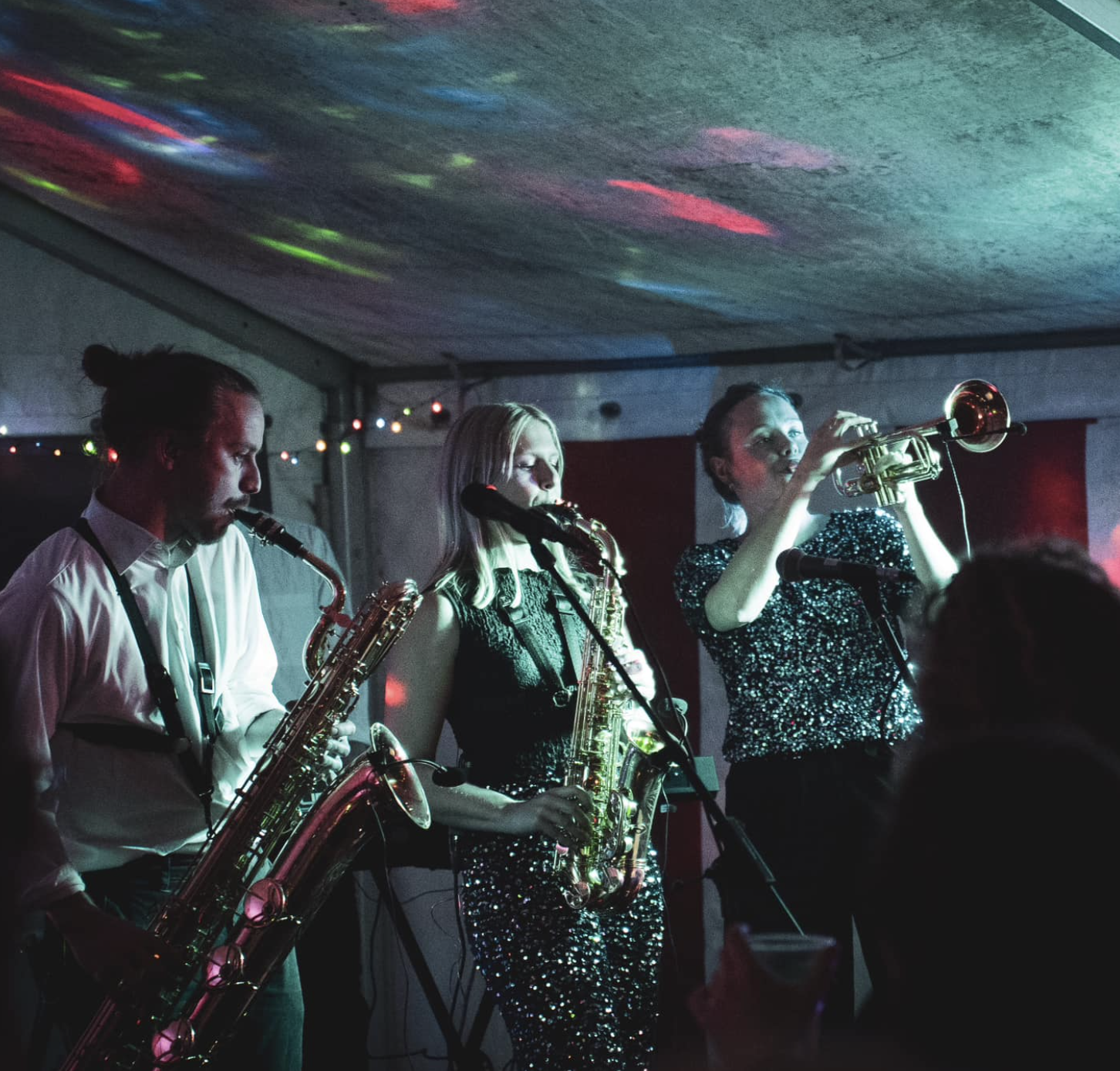 Liveband spiller på intim scene med farvet loftsbelysning og publikum tæt på, hvilket skaber en energisk og stemningsfuld koncertatmosfære